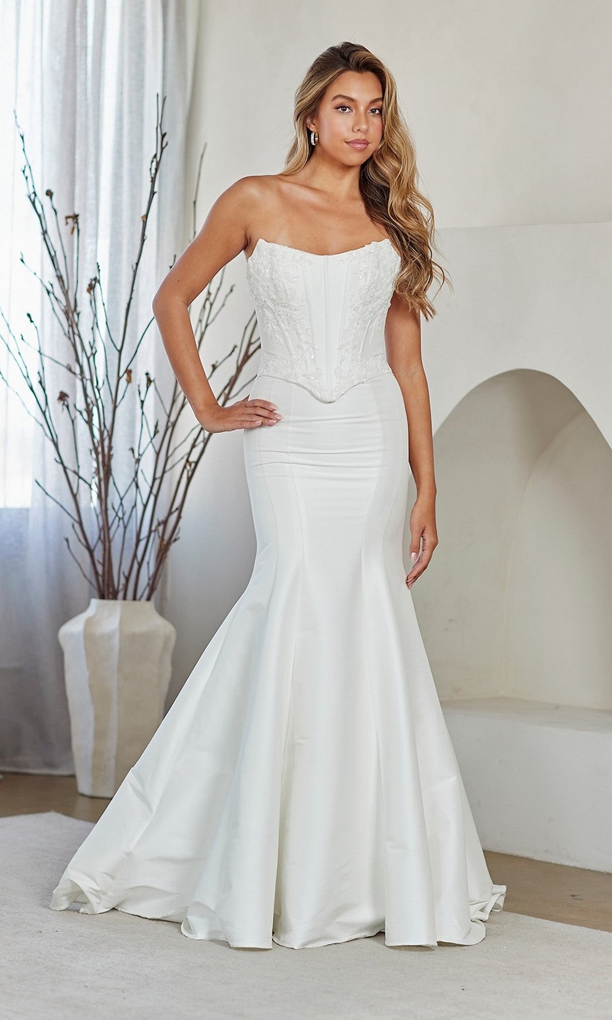 A woman stands indoors wearing the Amelia Couture 8080 long white prom dress, which has a strapless mermaid silhouette with an embellished corset bodice and fit-and-flare skirt. A dried branch arrangement and arched niche are visible in the background.